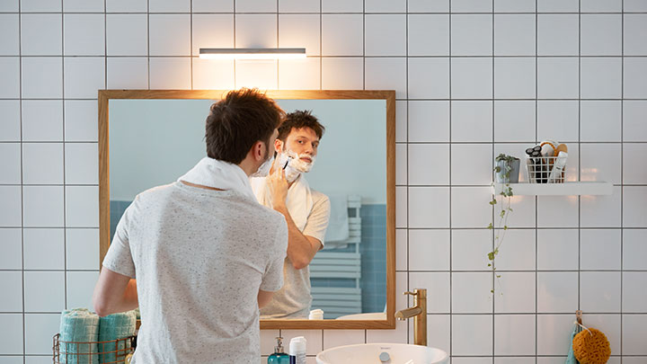 Homem a barbear-se em frente a um espelho de casa de banho iluminado por um LED da Philips montado na parede