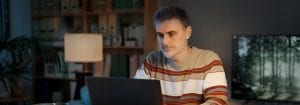 Um homem com uma camisola às riscas senta-se à secretária e olha para o computador portátil que tem à sua frente